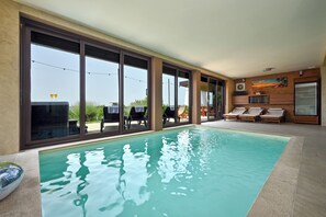 A modern indoor swimming pool with clear glass walls, lounge chairs, and a wooden ceiling. - Modern villa for 5 with indoor pool, jacuzzi, sauna, and barbecue (Marija Bistrica)