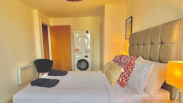 Ett hotellrum med en säng, ett nattduksbord med en lampa, en stol, en tvättmaskin och en dörr.