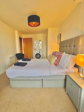 A hotel room with a bed, a bedside table with a lamp, a chair, a washing machine, and a door. - Stunning Shard Views (London)