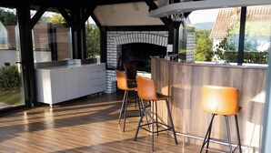 An outdoor kitchen with a brick fireplace, wooden bar counter, and two bar stools. - Luxury villa with jacuzzi, sauna and pool surrounded by vineyards (Požega)