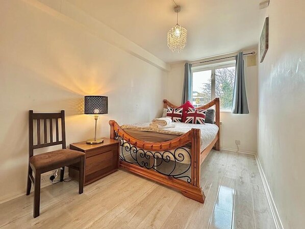 A bedroom with a wooden bed, a bedside table with a lamp, a chair, and a window with curtains. - Right Opposite Whitechapel Station (London)