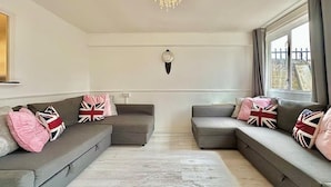 A modern living room with two grey sofas, each adorned with British flag pillows. A chandelier hangs from the ceiling, and a clock is mounted on the wall. - Right Opposite Whitechapel Station (London)