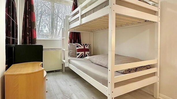A bunk bed room with a window, a radiator, and a small wooden stool. - Right Opposite Whitechapel Station (London)