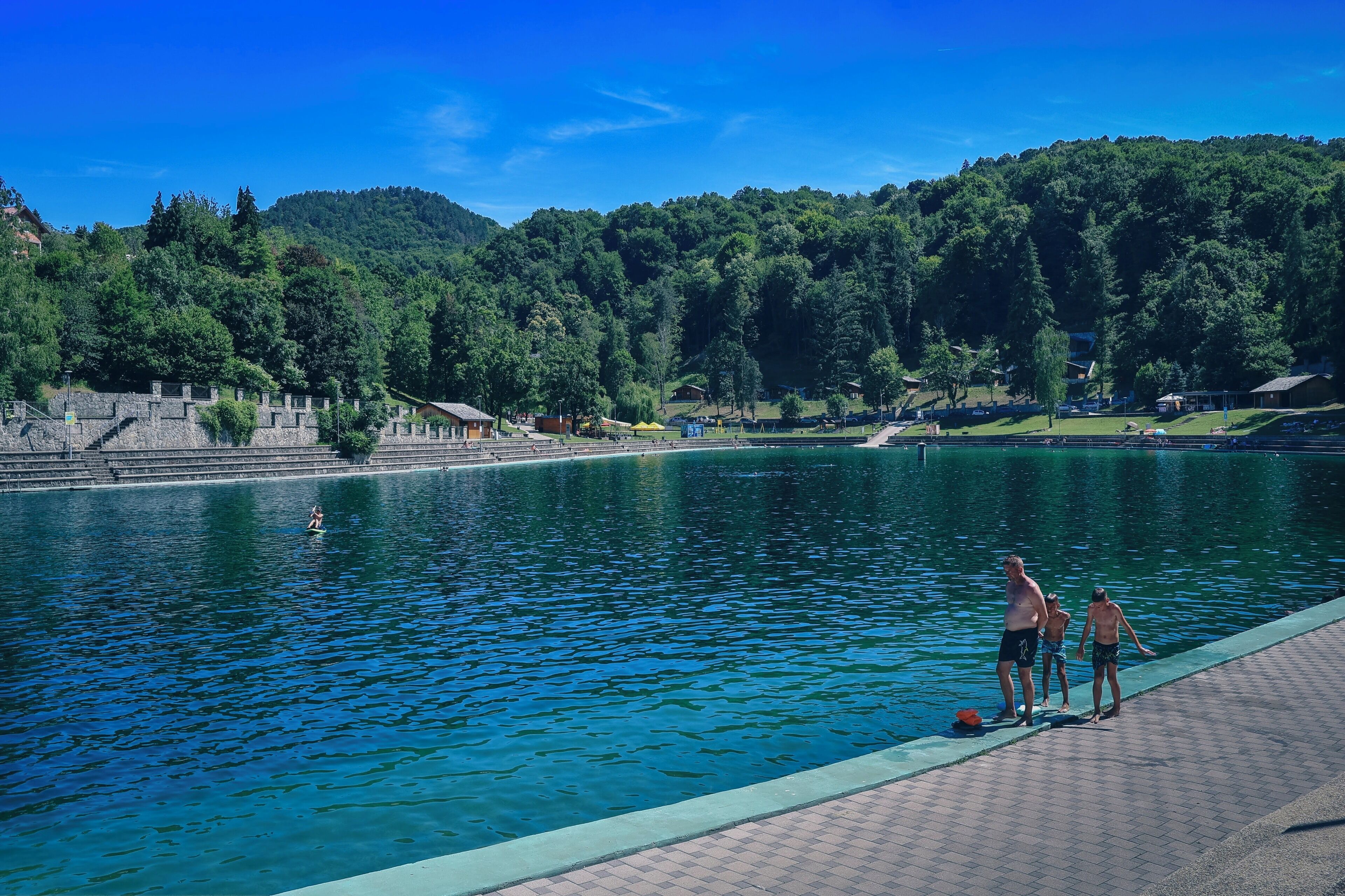 A swimming pool with a stone wall and steps leading into the water, surrounded by a forested hillside.