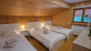 Une chambre d’hôtel avec des boiseries, trois lits, des lampes de chevet, un radiateur et une fenêtre donnant sur un paysage verdoyant.