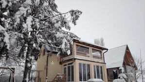 Een tweelaags huis met een met sneeuw bedekte dak en ramen, omringd door met sneeuw bedekte bomen en een heldere lucht.