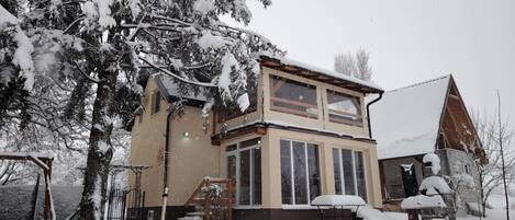 A two-story house with a snow-covered roof and windows, surrounded by snow-covered trees and a clear sky.