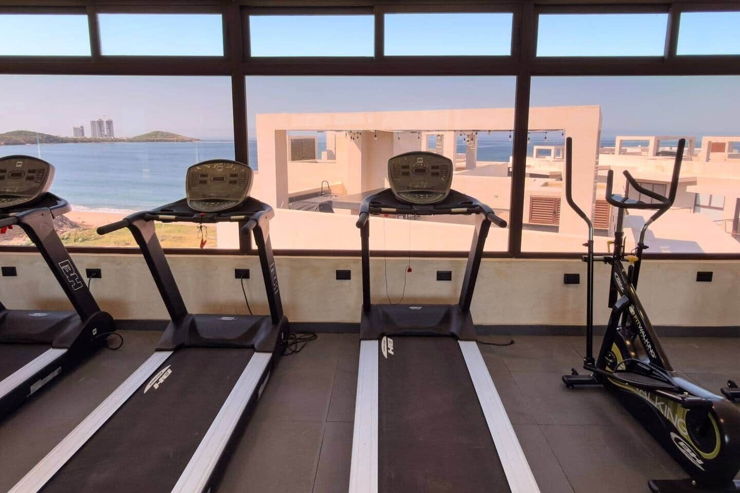 Ein Fitnessstudio mit Laufbändern und einem Ergometer mit Blick auf Strand und Gebäude.