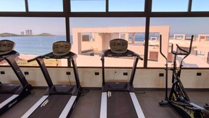 Ein Fitnessstudio mit Laufbändern und einem Ergometer mit Blick auf Strand und Gebäude.