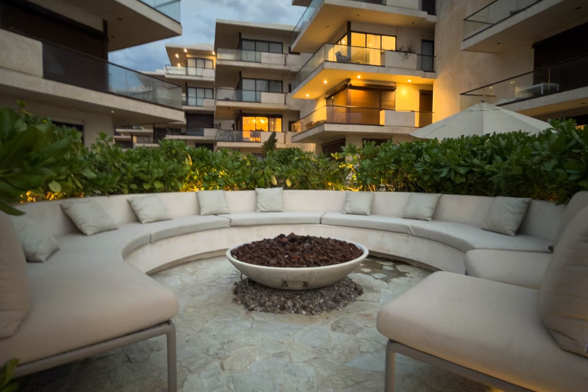 A modern outdoor seating area with a curved sofa and a central fire pit, surrounded by lush greenery and a multi-story building in the background.
