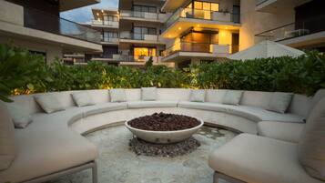 A modern outdoor seating area with a curved sofa and a central fire pit, surrounded by lush greenery and a multi-story building in the background.