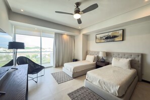 Une chambre d’hôtel comprenant un lit, deux tables de chevet, un bureau, une chaise, un ventilateur de plafond et une grande fenêtre avec des rideaux.
