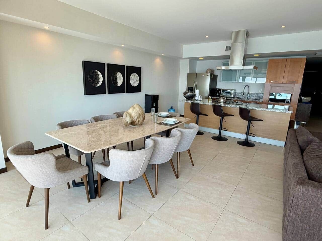 Een moderne keuken met een centraal eiland, een eetgedeelte met een grote tafel en stoelen, en een zithoek met een bank.