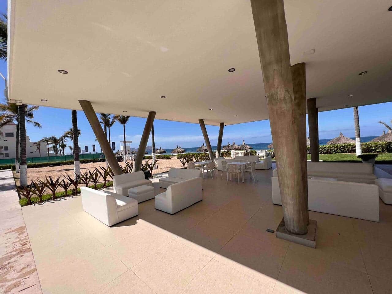 Ein überdachter Außenbereich mit weißen Sofas und Tischen, Palmen und Blick auf Strand und Meer.