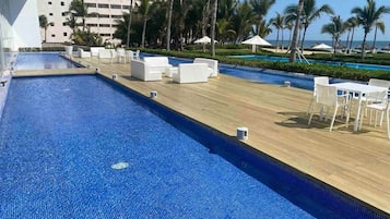 A pool area with lounge chairs and umbrellas, palm trees, and a building in the background.