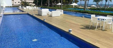 A pool area with lounge chairs and umbrellas, palm trees, and a building in the background.