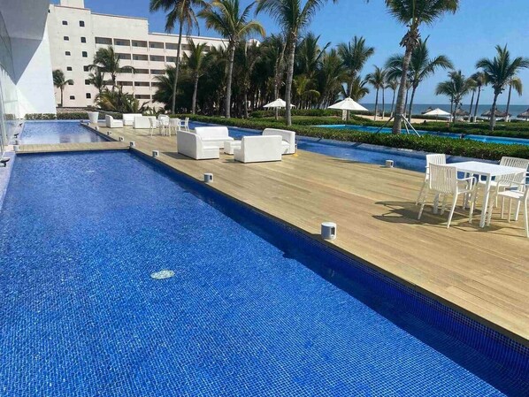 Zona de piscina con sillones de descanso y sombrillas, palmeras y un edificio al fondo.