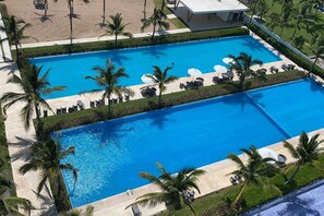 Une grande piscine rectangulaire entourée d’une terrasse, de palmiers et de chaises longues.