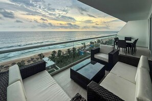 Un balcón con vistas a la playa y al océano, amoblado con sofás de mimbre y una mesa de centro de vidrio.