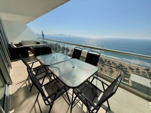 Un balcon avec une table et des chaises en verre, donnant sur une plage et un pont.