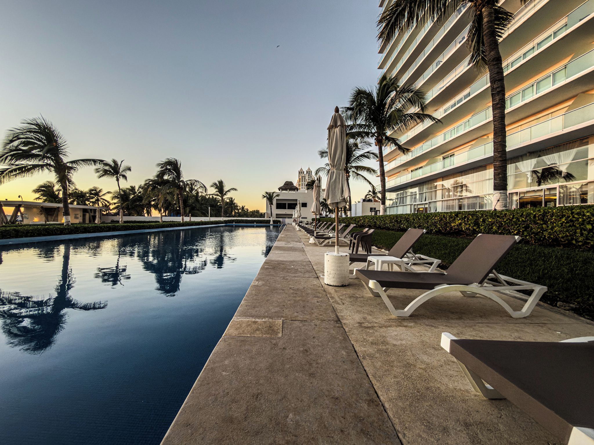 A swimming pool with lounge chairs and palm trees, surrounded by a hotel building.