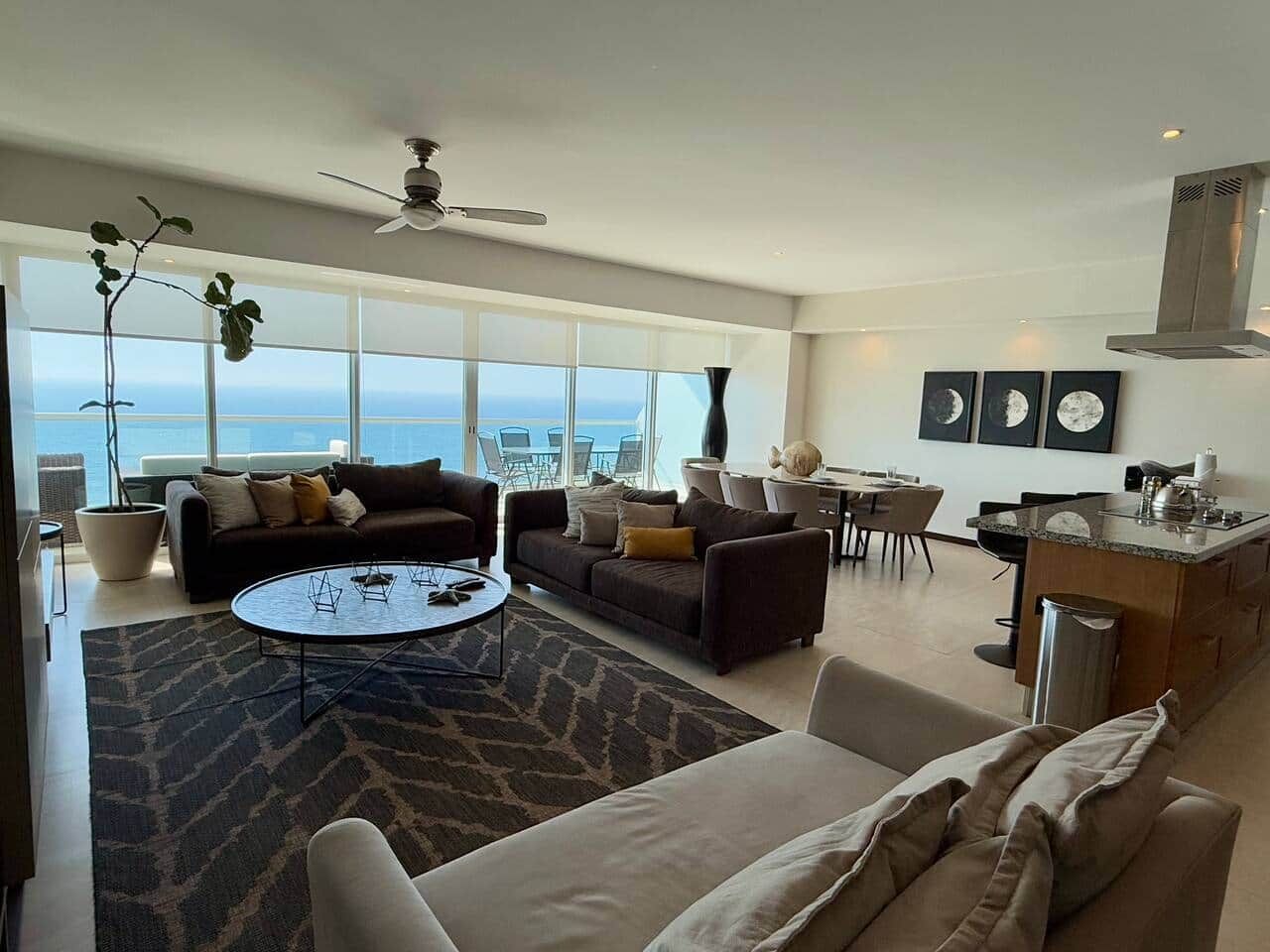 A modern living room with a sofa, coffee table, and dining area, large windows with a view, and a kitchen with a countertop and sink.