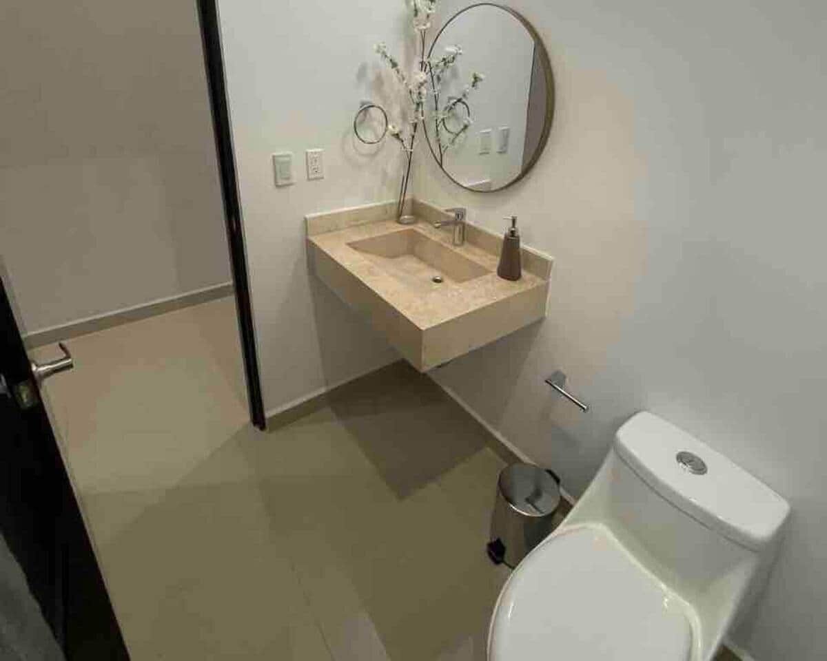 Un baño con un lavamanos color beige, un espejo, un inodoro y un cubo de basura.