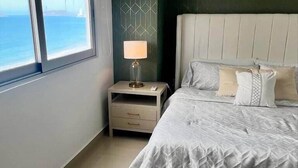 Een slaapkamer met een groot bed, een nachtkastje, een lamp en uitzicht op de zee door een raam.