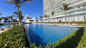 A swimming pool with lounge chairs, palm trees, and a modern hotel building in the background. - Peninsula Mazatlan Floor 27: Exclusive 3-Bedroom Oceanfront (Mazatlan)
