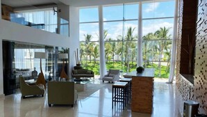 A spacious lobby with a high ceiling, large windows offering a view of palm trees, and a mix of seating arrangements including sofas and chairs. - Peninsula Mazatlan Floor 27: Exclusive 3-Bedroom Oceanfront (Mazatlan)