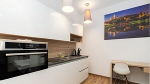 Cozinha moderna com armários brancos, forno embutido e bancada de madeira. Há uma escrivaninha e uma cadeira, e um quadro com vista para a cidade na parede.