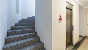 Escada que dá acesso a um elevador em um corredor com paredes brancas e um alarme de incêndio na parede.