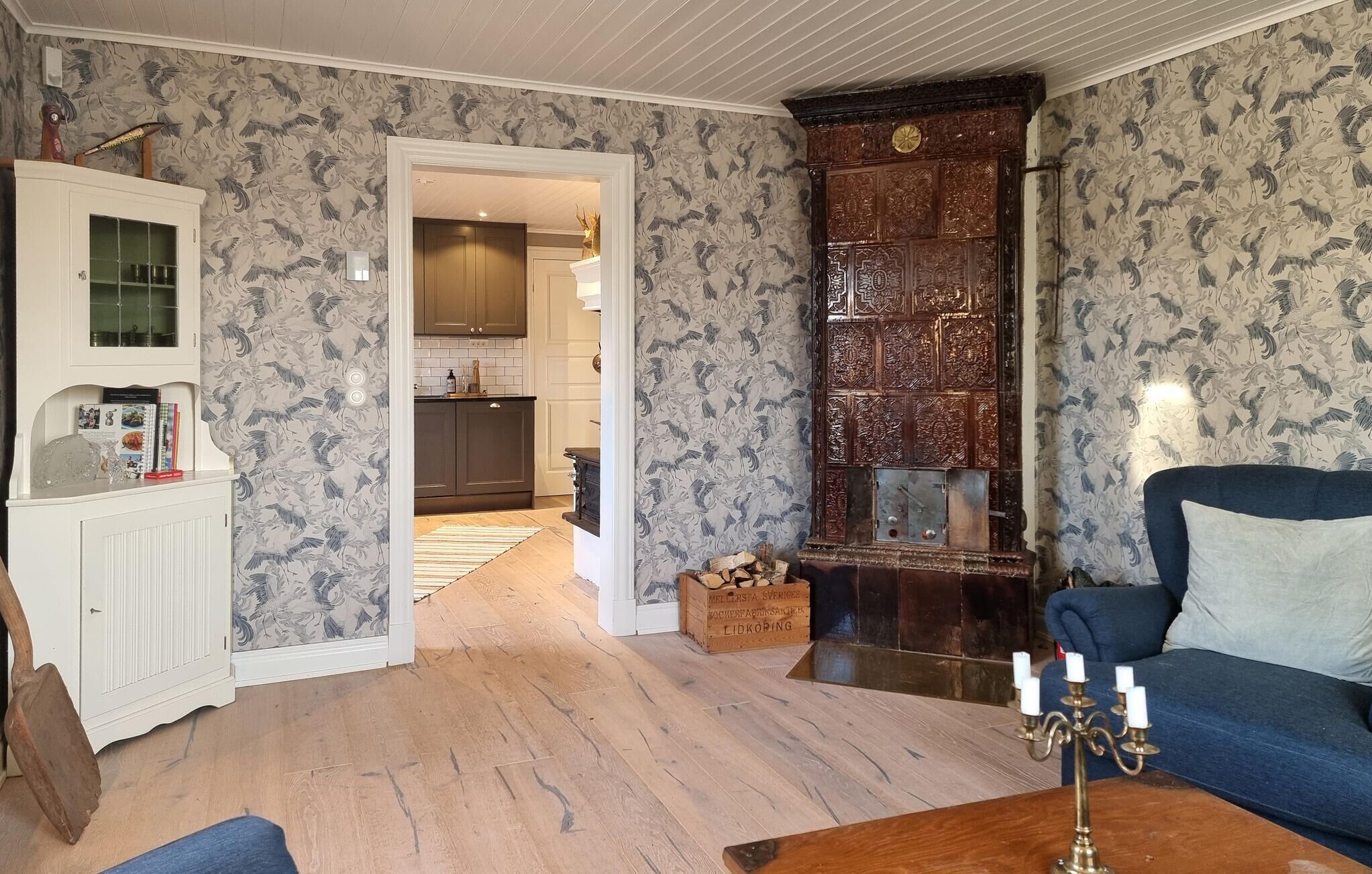 Une pièce avec un plancher en bois, un canapé bleu, un meuble blanc aux portes vitrées et un papier peint à motifs.