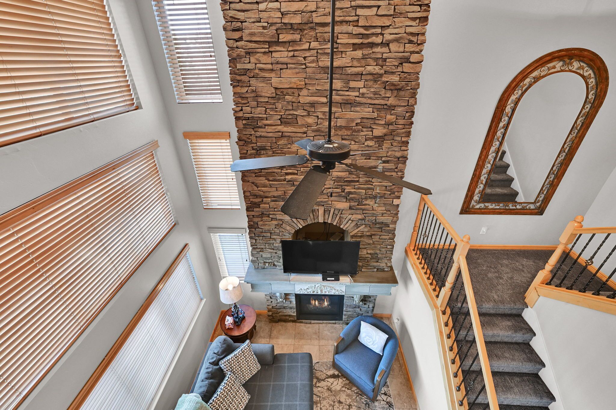 A living room with a stone fireplace, a ceiling fan, and a staircase with wooden railings.