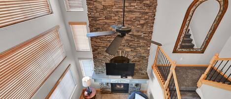 A living room with a stone fireplace, a ceiling fan, and a staircase with wooden railings.