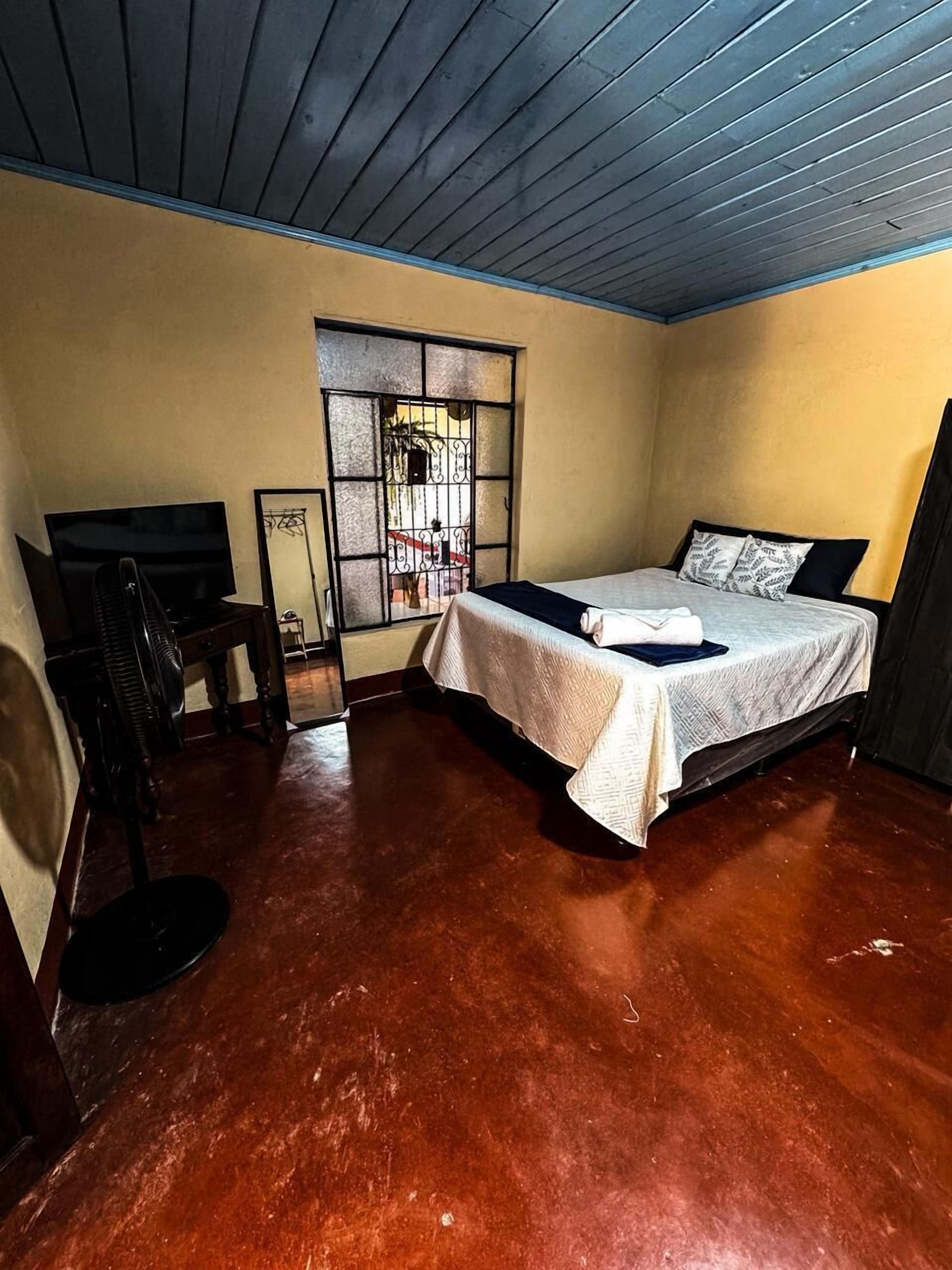 A bedroom with a bed, a wooden ceiling, a window with bars, and a small table with a chair.