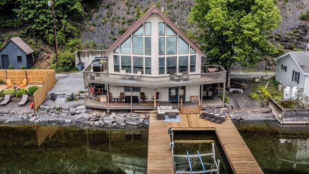 Een groot, modern huis met een opvallende glazen gevel, een houten terras en een steiger die uitsteekt in kalm water.