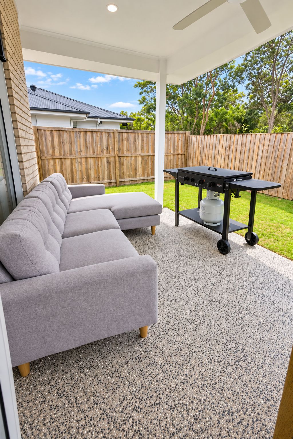 Un patio couvert avec un canapé d’angle gris, un barbecue noir et une clôture en bois en arrière-plan.