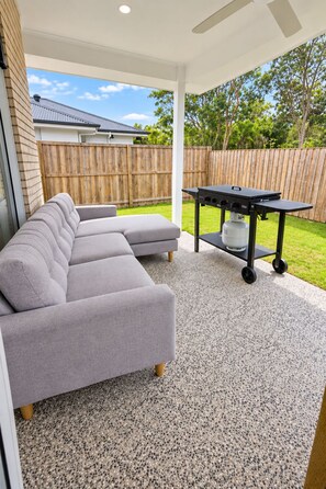 A covered patio area with a grey sectional sofa, a black grill station, and a wooden fence in the background. - Serene 4-bedroom house with AC in charming Park Ridge (Park Ridge)