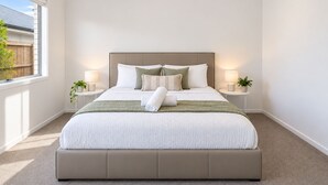 Un dormitorio con una cama grande, dos mesitas de noche con lámparas, una ventana con vistas a una valla y una planta en cada mesita.