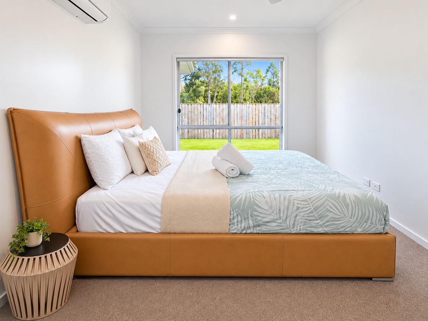 A modern bedroom with a large bed, a potted plant, and a view of a fenced garden through a sliding door.