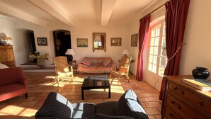 Uma sala de estar acolhedora com um sofá, poltronas, uma mesa de centro e um armário de madeira. Há uma janela com cortinas, uma porta e um abajur sobre uma mesinha lateral.