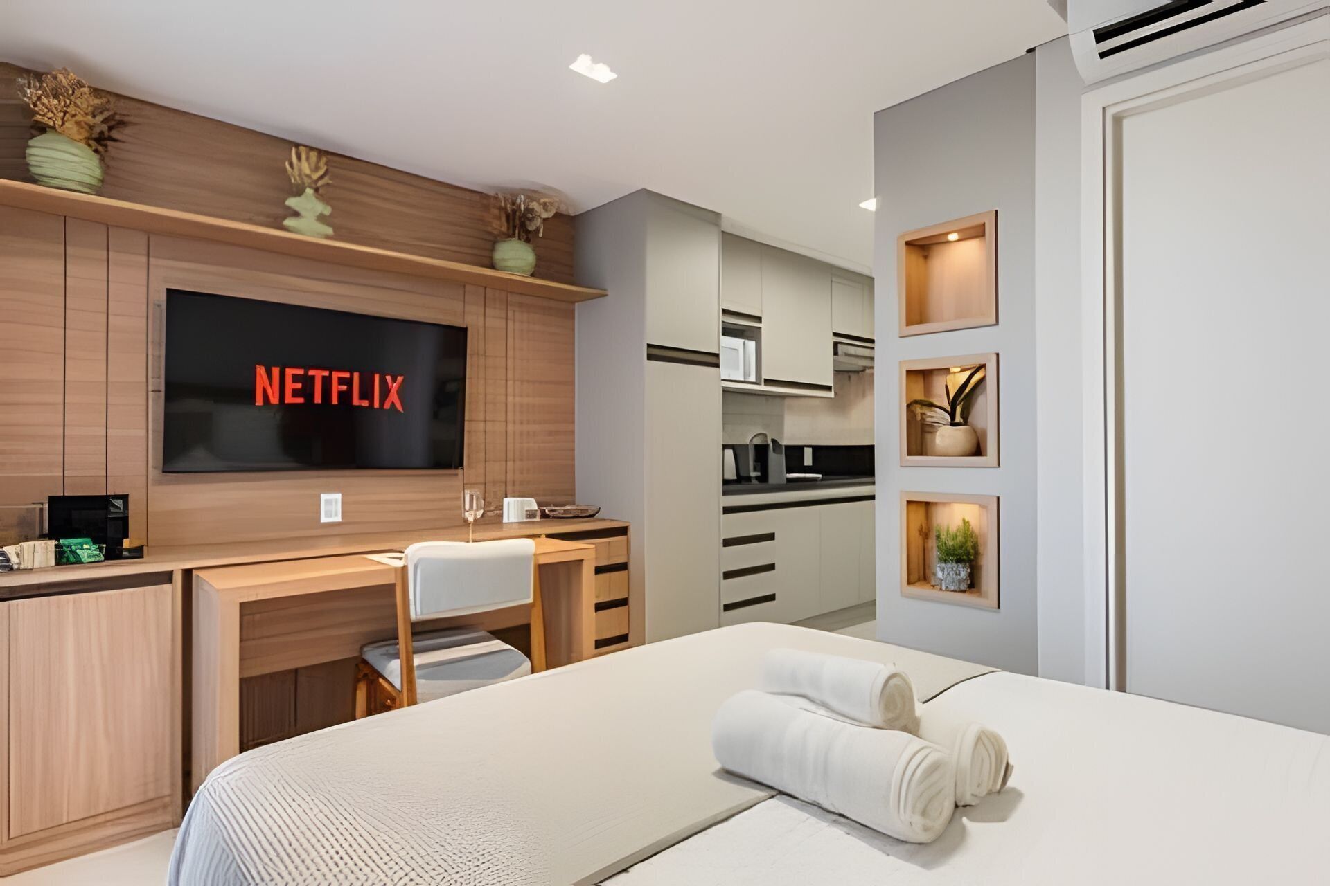 A modern hotel room with a large bed, a desk with a computer, a TV displaying 'NETFLIX,' and a kitchenette.