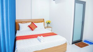 A neatly made bed with a wooden headboard, a red and white pillow, a white blanket, and a rolled towel on the bed. There is a small nightstand with a vase of yellow flowers beside the bed. - Gia Bao Hotel Thu Duc (Ho Chi Minh City)