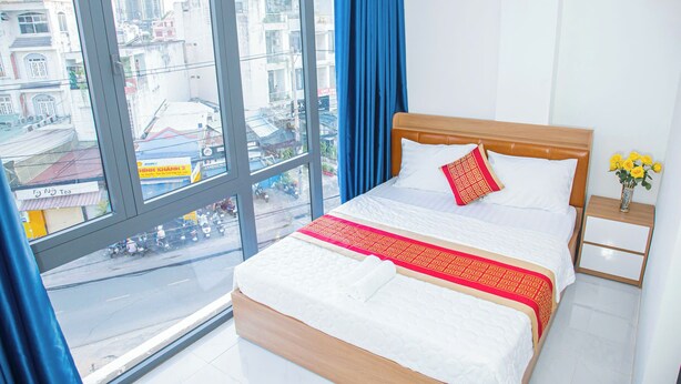 A hotel room with a large window, a bed with white bedding and a red and gold throw, a wooden nightstand with a vase of yellow flowers, and a view of a busy street and buildings. - Gia Bao Hotel Thu Duc (Ho Chi Minh City)