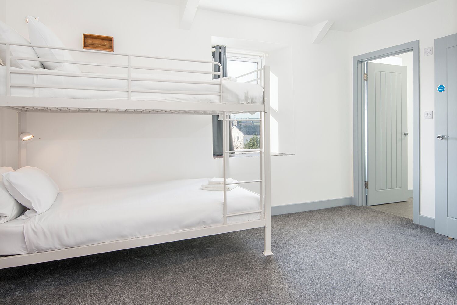 A bunk bed room with white bedding, a small wooden shelf, and a door leading to another room.
