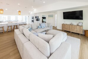 A modern living room with a large sectional sofa, a flat-screen TV, and a kitchen area in the background. - SEASIDE RETREAT - 8 bedroom semi-oceanfront with pool and ocean views (Virginia Beach)
