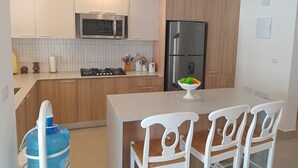 Eine moderne Küche mit Holzausstattung, einem Edelstahlkühlschrank, einer Mikrowelle und einem Gasherd. Ein Esstisch mit weißen Stühlen und eine Schüssel Obst stehen darauf.