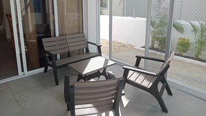 Eine Terrassenfläche mit einer Holzbank und zwei Stühlen, einem Tisch und einer Glastür, die zu einem überdachten Außenbereich führt.