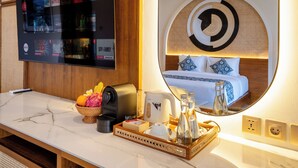 Ein Hotelzimmer mit einem großen Fernseher, der verschiedene Sender zeigt, einem Bett mit einem runden Spiegel darüber sowie einem Tablett mit Toilettenartikeln und einer Kaffeemaschine auf der Ablagefläche.